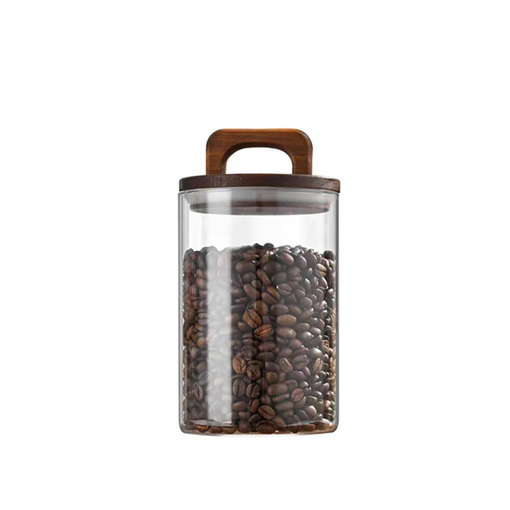 A transparent glass jar filled with coffee beans, featuring a cylindrical shape and a dark wooden lid with a built-in handle, set against a plain white background.