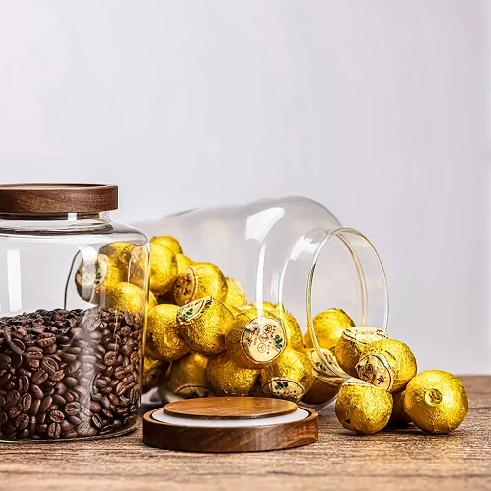 A set of transparent glass spice jars with wooden lids is displayed on a wooden surface. One jar is filled with dark roasted coffee beans, while another has been tipped over, spilling gold-wrapped chocolates. The wooden lids, featuring a silicone gasket for an airtight seal, are placed beside the jars. The scene is set against a neutral background, emphasizing the jars’ elegant and functional design for storing spices, coffee, or treats.