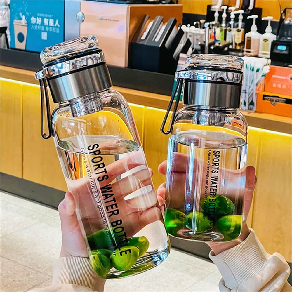 Two glass sports water bottles with stainless steel leakproof lids and built-in filters, filled with water and green limes, are being held by two people in a modern café setting. The transparent design showcases the infused water, emphasizing its functionality for fresh and healthy hydration. The background features a sleek counter with beverage accessories.