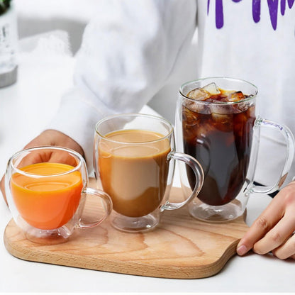 Three double-wall glass tumblers filled with orange juice, coffee, and iced coffee sit on a wooden tray, held by a person in a white sweatshirt.