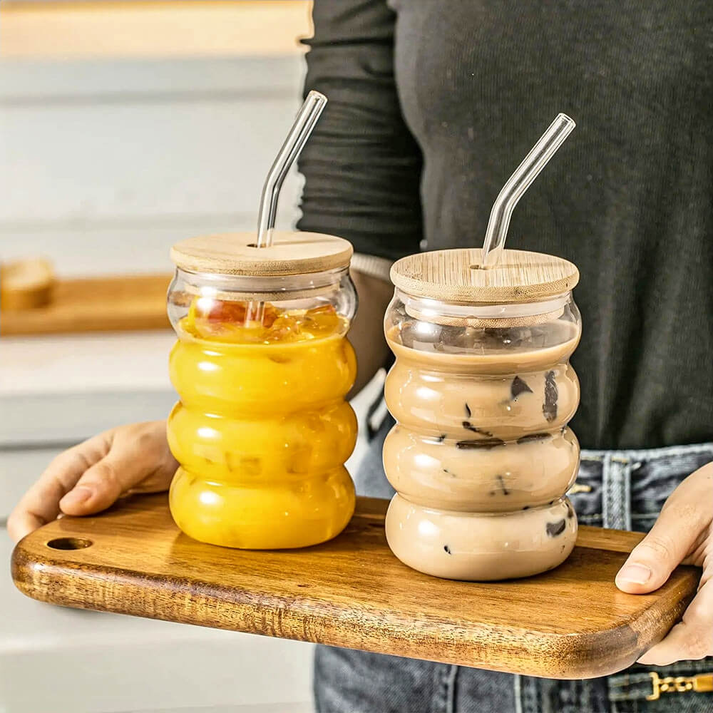Two wavy glass tumbler with bamboo lid designs, each with a reusable glass straw, filled with orange juice and iced coffee, placed on a wooden serving tray.