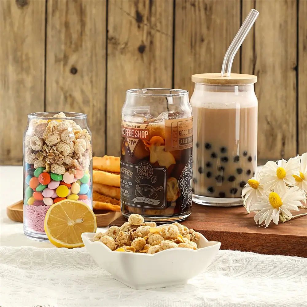 Glass tumbler with lid and straw holding iced milk tea, shown with coffee glass, cereal jar, snacks, and flowers on a wooden table.