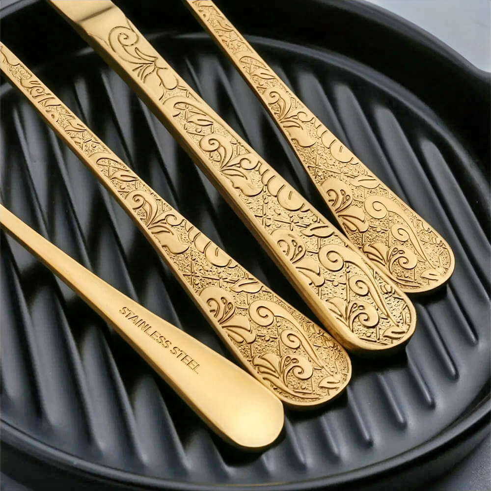 Close-up of gold silverware with intricate patterns on handles, crafted from stainless steel, displayed on a black textured surface.