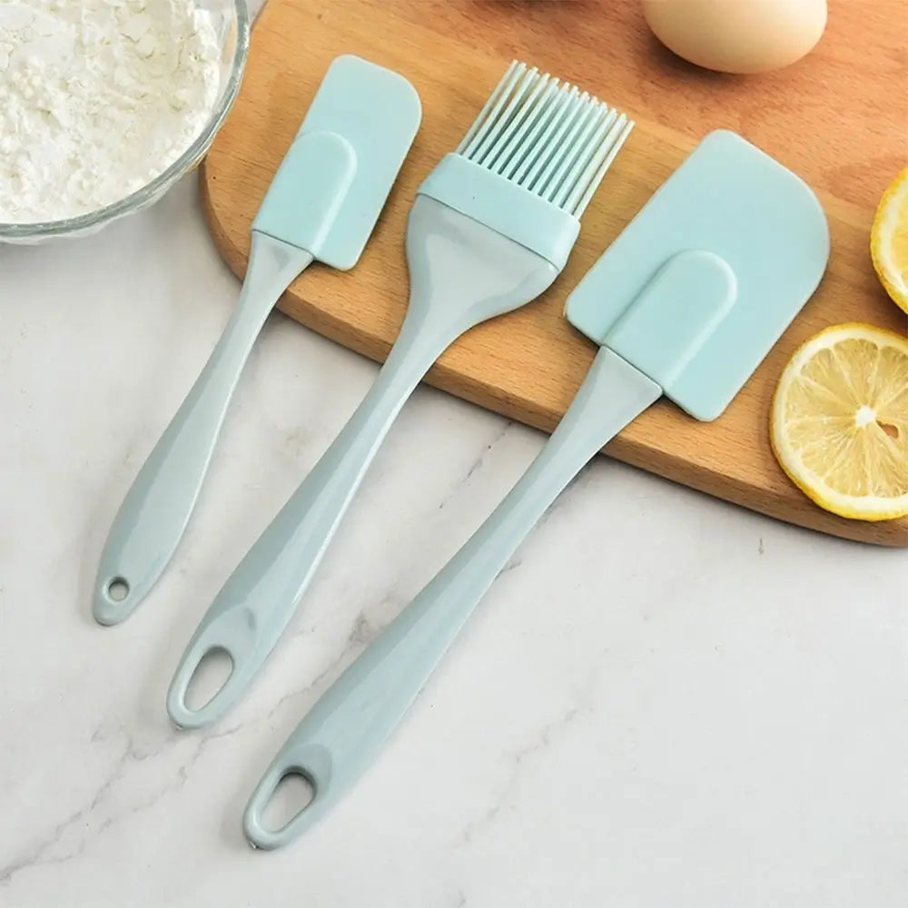 Green silicone kitchen utensils set with two spatulas and a pastry brush displayed beside lemons and baking ingredients.