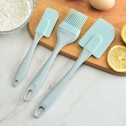 Green silicone kitchen utensils set with two spatulas and a pastry brush displayed beside lemons and baking ingredients.