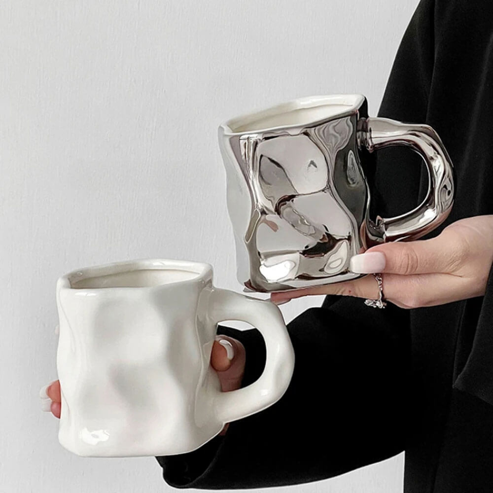 Two hand-glazed coffee mugs, one in a sleek white finish and the other in a reflective metallic finish, both featuring a unique crumpled texture. Each mug has a sturdy ergonomic handle and a modern design, perfect for adding a stylish touch to your coffee or tea experience.