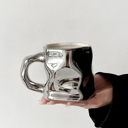 Hand holding a shiny, crumpled-design hand-glazed stoneware mug with a 16 oz capacity. The mug features a glossy metallic finish, a sturdy ergonomic handle, and a contemporary artistic shape, showcasing its unique craftsmanship.