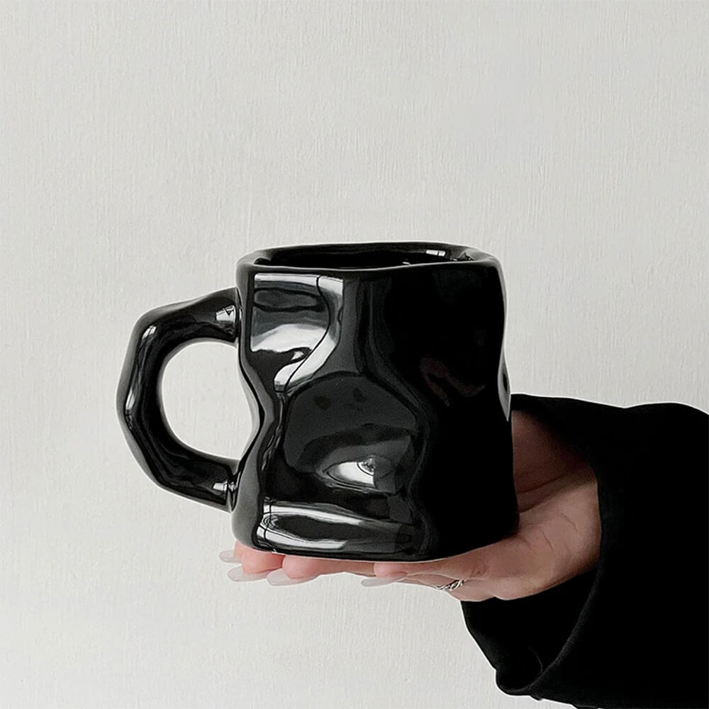 Hand holding a sleek black hand-glazed stoneware mug with a modern crumpled design. The mug features a glossy finish, a sturdy ergonomic handle, and a 16 oz capacity, showcasing its contemporary craftsmanship and unique style.