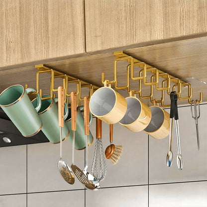 Gold-toned IKEA cabinet rack mounted under a wooden shelf, featuring multiple hooks neatly holding green and yellow mugs, utensils, and kitchen tools for a stylish and organized kitchen setup.