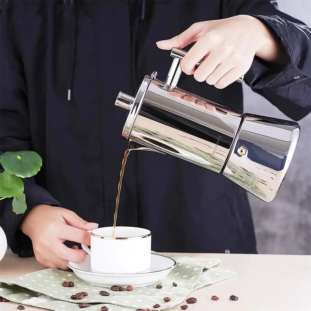 Person pouring coffee from a stainless steel induction stovetop espresso maker into a white cup on a saucer.