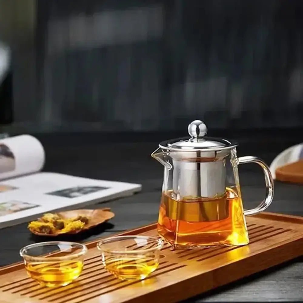 A glass teapot with stainless steel infuser and lid on a wooden tray, filled with amber tea, beside two small glass cups.