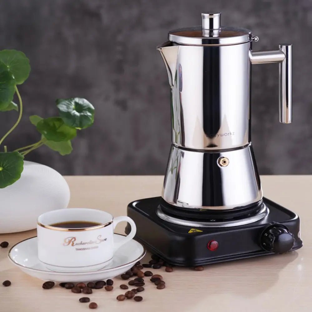 Stainless steel stovetop espresso maker placed on an electric burner beside a cup of coffee and scattered coffee beans.