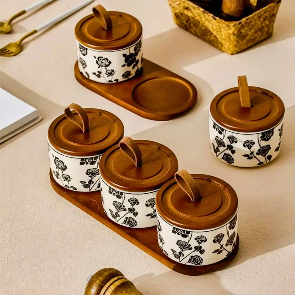 A floral ceramic kitchen canister sets collection featuring multiple white containers with black rose patterns, each topped with a wooden lid and leather loop handle. The sets are displayed on matching wooden trays.