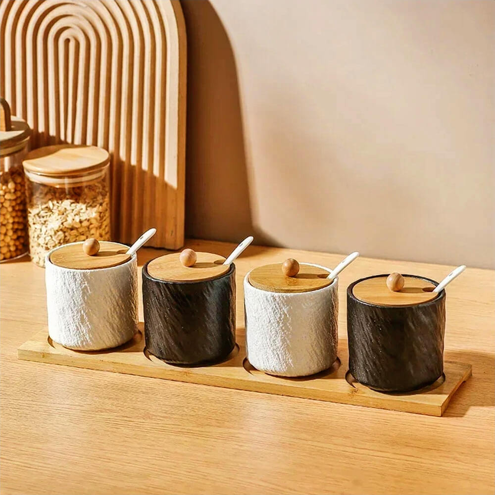 Four ceramic jars with a textured finish, alternating in black and white, are arranged on a rectangular bamboo tray. Each jar has a bamboo lid with a wooden knob and a small white spoon resting in a slot on the lid. The setup is placed on a wooden surface with warm lighting, accompanied by a wooden arch and jars filled with grains in the background.