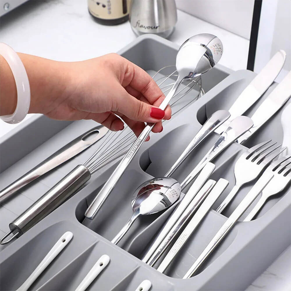 A gray kitchen drawer organizer holding neatly arranged cutlery, utensils, and a whisk. A hand is picking up a spoon, demonstrating easy access and organization.