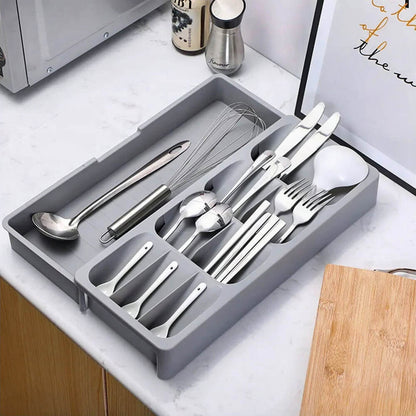 A gray kitchen organizer drawer with an expandable design, neatly storing cutlery, utensils, and kitchen tools. Placed on a white countertop alongside a wooden cutting board, framed wall art, salt and pepper shakers, and a whisk for a well-organized kitchen setup.