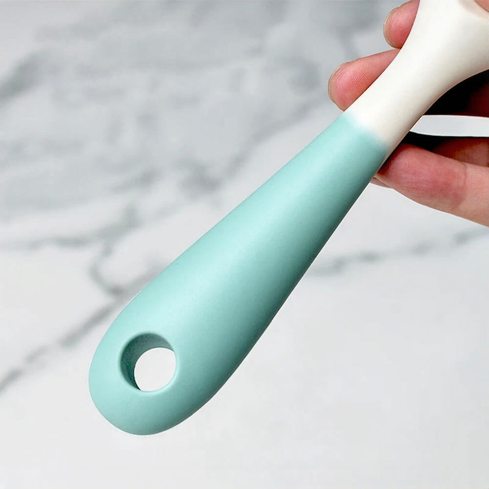 A close-up of a kitchen silicone utensil set handle, featuring a smooth, ergonomic grip in a pastel green shade with a white transition near the top. The handle has a convenient hanging hole for easy storage. The background is a marble countertop, providing a stylish and modern kitchen aesthetic.