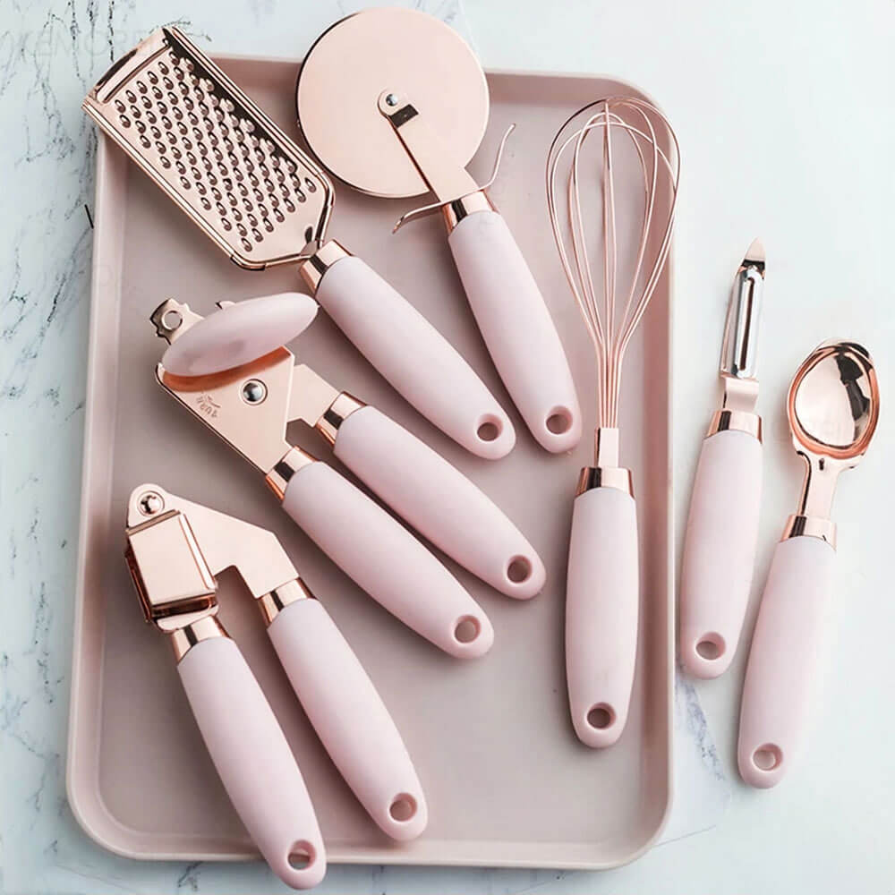 A stainless kitchen tool set featuring rose gold accents and soft pink handles, including a whisk, grater, pizza cutter, garlic press, ice cream scoop, can opener, and peeler, elegantly arranged on a light pink tray for a modern and stylish look.