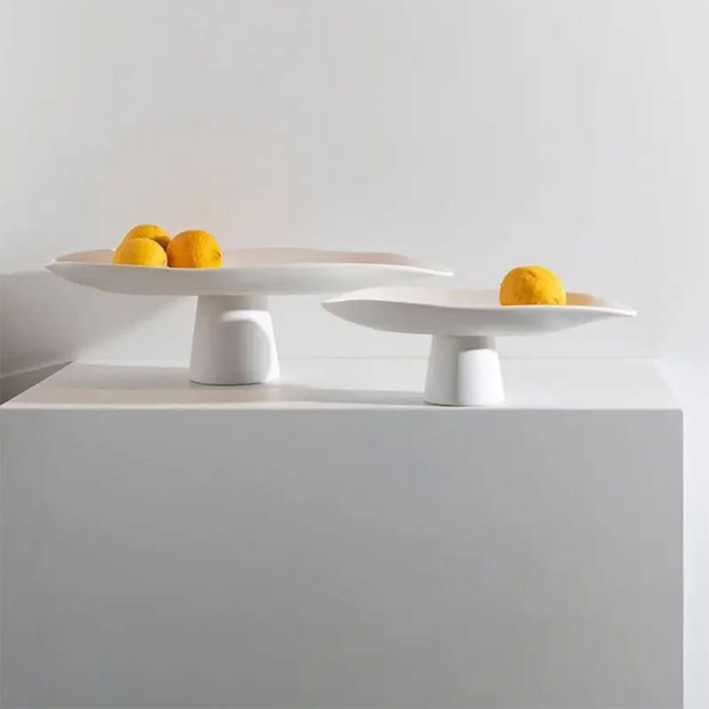 Two minimalist white ceramic fruit bowls, including a large ceramic fruit bowl with a wide, flat surface and a sturdy pedestal base. The larger bowl contains several vibrant oranges, while the smaller bowl holds a single orange, creating an elegant and modern display against a neutral background.