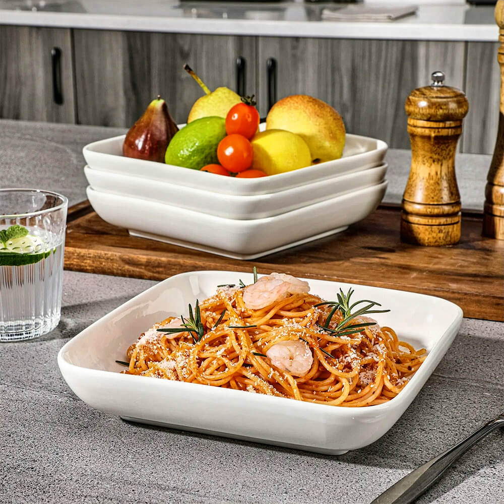 A set of large serving bowls in white porcelain, displayed on a kitchen counter. One bowl contains shrimp pasta, while a stacked set holds assorted fruits. A wooden cutting board, salt and pepper grinders, a glass of water with lime, and a fork are arranged nearby.