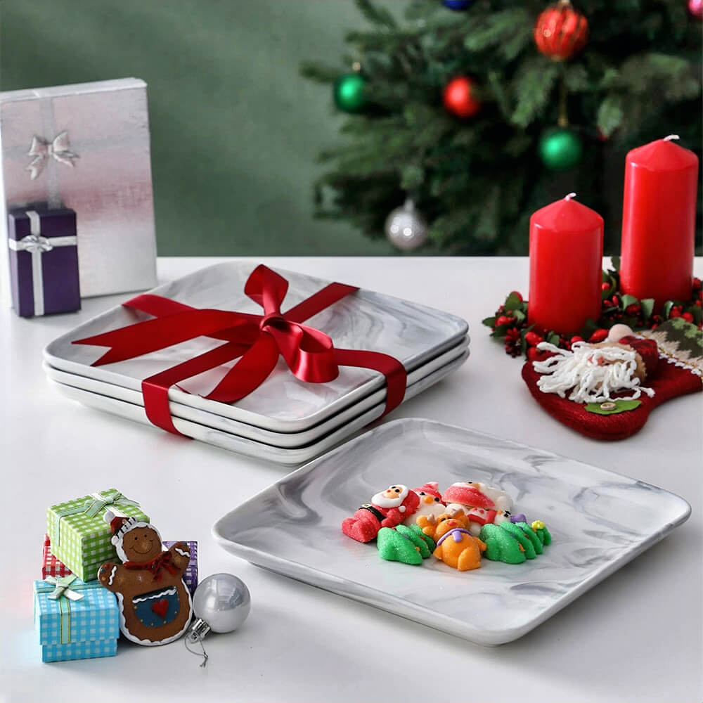 A stack of marble-style square serving plates tied with a red ribbon, placed on a festive table with Christmas decorations, gifts, and red candles. One large serving plate is displayed with colorful holiday-themed treats. A decorated Christmas tree is visible in the background.