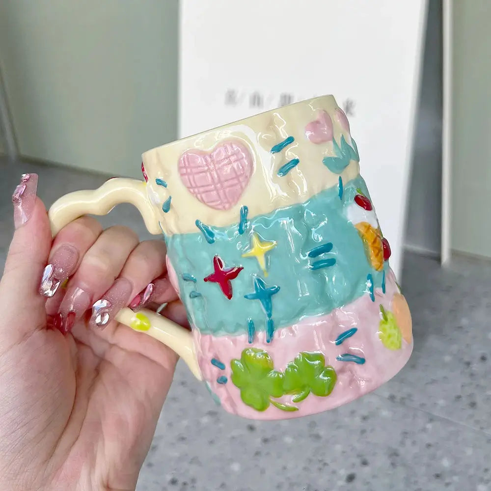 Hand holding a colorful latte ceramic mug with a heart-shaped handle, raised motifs, and a glossy handcrafted finish.
