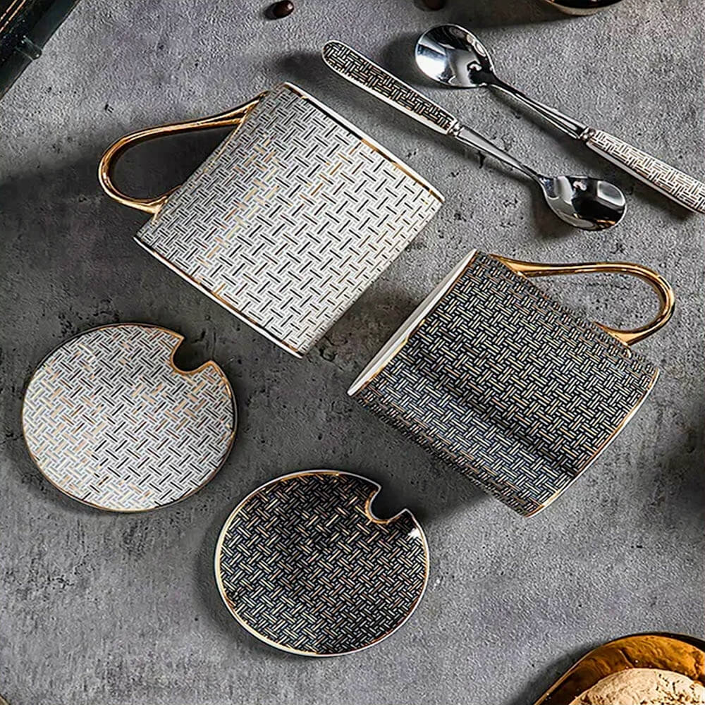 A set of ceramic coffee mugs with their matching lids and spoons arranged on a textured surface. The mugs feature intricate geometric patterns in black and white with gold accents, including gold handles for a luxurious touch. The lids are designed to perfectly fit the mugs, with a small opening for spoons, making them both stylish and practical for preserving warmth.