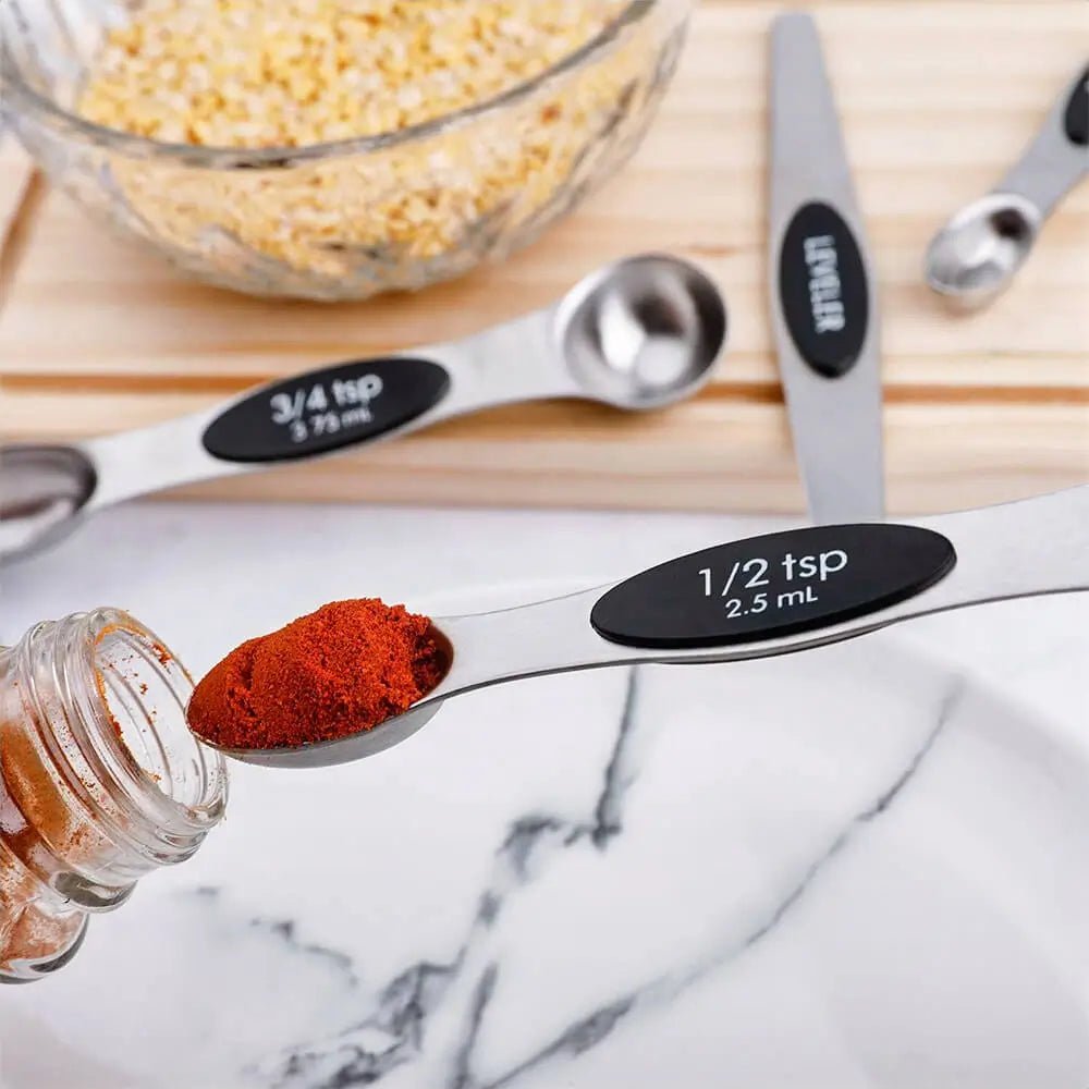 Measuring spoons with labeled capacities on a marble surface, next to a jar of red powder and a bowl of yellow seeds.