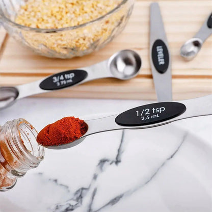 Measuring spoons with labeled capacities on a marble surface, next to a jar of red powder and a bowl of yellow seeds.