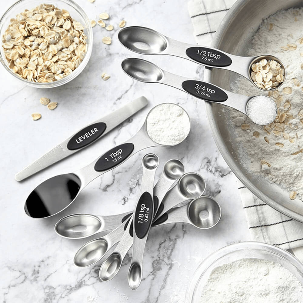 A set of stainless steel magnetic measuring spoons displayed on a marble surface alongside ingredients like oats, flour, and sugar. The spoons include a leveler tool and feature dual-sided designs for accurate measurement of liquids and dry ingredients.