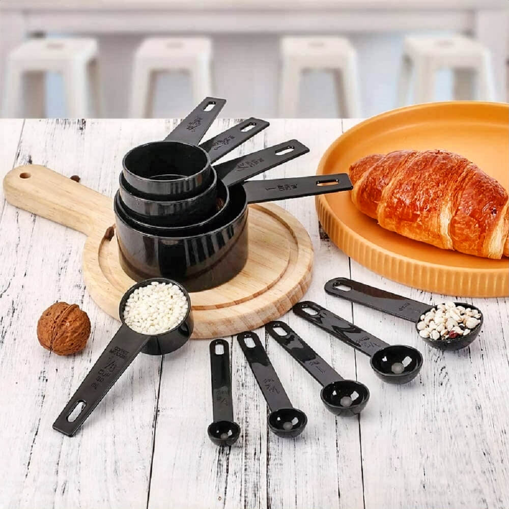 Black measuring cups and measuring spoons filled with ingredients on a wooden table next to a croissant and cutting boards.