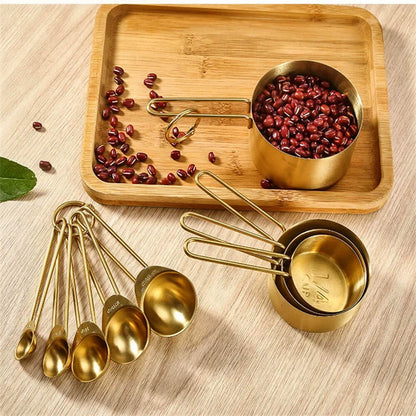 A beautifully arranged set of measuring cups and spoons stainless steel with a luxurious gold finish displayed on a wooden tray. The measuring cups are filled with red beans, showcasing their functionality, while the spoons are neatly laid out nearby. The warm wood tones complement the elegant gold design, emphasizing a blend of style and practicality for kitchen use.