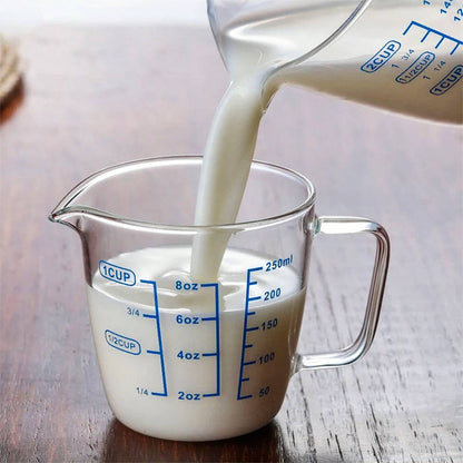 A 1-cup glass measuring cup being filled with milk from a 2-cup glass measuring cup. Both feature blue measurement markings for ounces, cups, and milliliters, with curved spouts and sturdy handles, placed on a wooden surface.