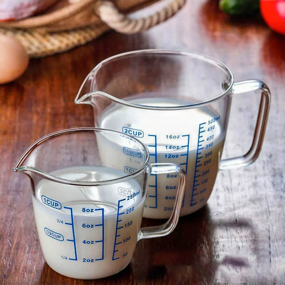 Two clear glass measuring cups on a wooden surface, one with a 1-cup capacity and the other with a 2-cup capacity. Both have blue measurement markings for ounces, cups, and milliliters, along with a curved spout and sturdy handle.