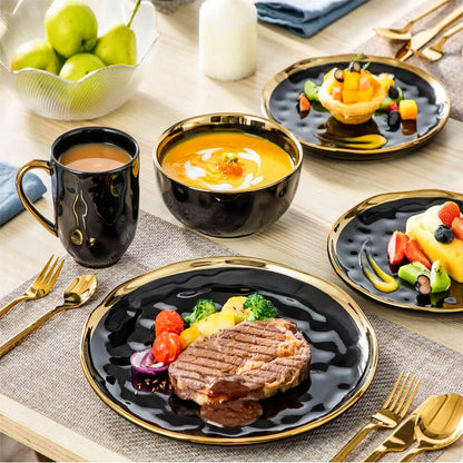 A Mendocino stoneware dinnerware set, 12-piece, displayed in an elegant dining arrangement. The set features glossy black plates and bowls with gold rims and accents, paired with matching black mugs with gold handles. A plate holds a grilled steak with roasted vegetables, while another contains a vibrant dessert of fresh fruits. A bowl is filled with creamy soup, and gold cutlery complements the sophisticated look. The table includes additional elements such as a bowl of green apples, a white candle, and te