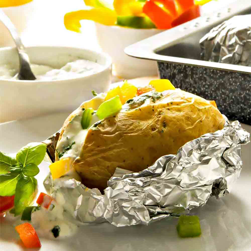 Baked potato wrapped in foil, topped with creamy sauce and colorful chopped vegetables, placed on a white plate—representing a traditional alternative to using microwave bags for potatoes.