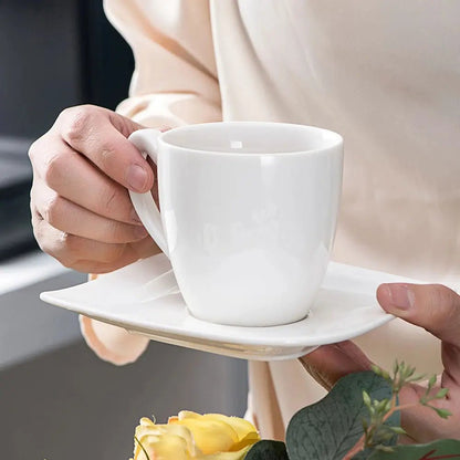 Modern everyday dinnerware with a white ceramic cup placed on a square white saucer, held in hand near a bouquet of yellow flowers.