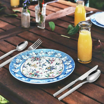 A beautifully arranged outdoor table featuring a modern flatware set made of 18/10 stainless steel. The utensils display a polished finish and textured handles, blending durability with a sleek, contemporary design. The setting includes a blue floral-patterned plate paired with a vibrant, embroidered napkin, all on a rustic wooden table. A small glass bottle of fresh orange juice adds a refreshing touch, while the natural greenery and delicate floral vases enhance the inviting and elegant atmosphere.