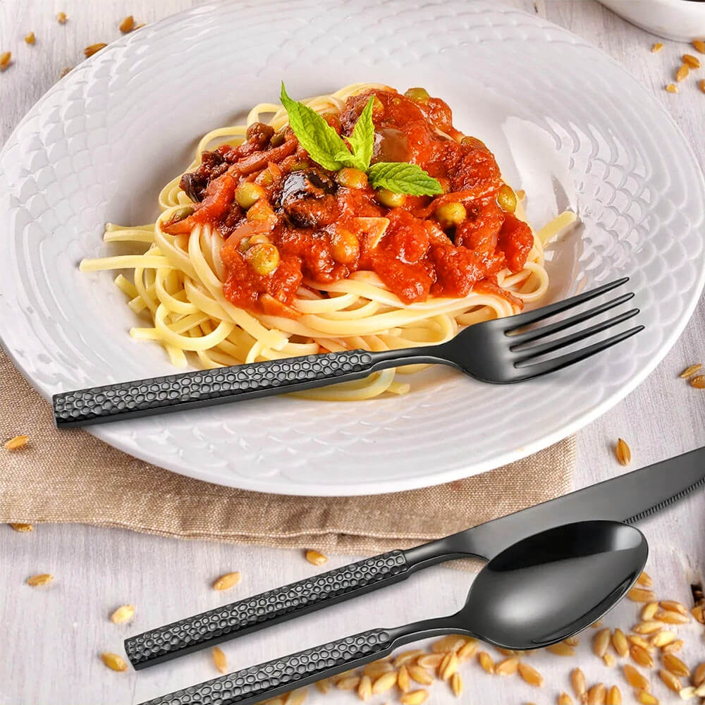 A stylish table setting featuring a modern flatware set for 12 in a matte black finish. The utensils showcase a textured handle design that adds a contemporary touch to the dining experience. A beautifully plated dish of spaghetti with rich tomato sauce, garnished with fresh mint, sits on a white patterned plate, resting on a neutral linen tablecloth. The sleek black flatware perfectly complements the vibrant food presentation, creating a modern and sophisticated dining atmosphere.