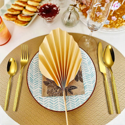 A beautifully styled dining table featuring a modern gold flatware set. The utensils have a textured handle design and a polished gold finish, complementing the elegant table setting. A fan-folded napkin sits atop a patterned plate with a floral-themed napkin, all placed on a gold charger plate. The scene is enhanced with champagne flutes, delicate vases, and an assortment of food, creating a luxurious and inviting atmosphere.