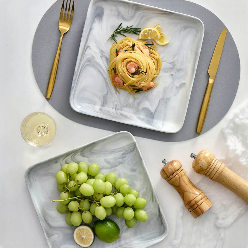 Two modern serving plates with a marble-style gray and white design on a dining table. One plate holds shrimp pasta garnished with lemon and rosemary, while the other contains green grapes and limes. The table setting includes gold cutlery, a glass of white wine, wooden salt and pepper grinders, and a gray placemat.