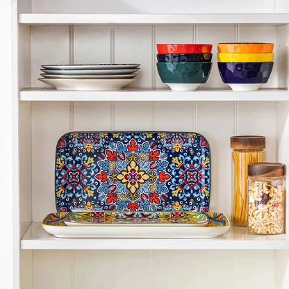 Modern serving plates with colorful floral and geometric patterns displayed on a kitchen shelf beside bowls, plates, and jars.