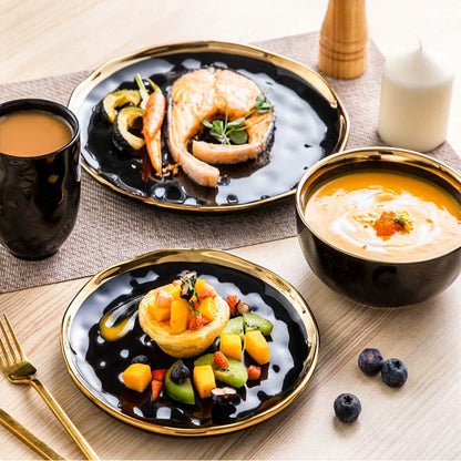 A modern stoneware dinnerware set arranged with an elegant meal presentation. The set features glossy black plates and bowls with gold accents on the rims, paired with a matching black mug with a gold handle. The plates showcase a variety of dishes, including a grilled salmon steak with roasted vegetables, a dessert tart topped with fresh mango, kiwi, and berries, and a creamy soup garnished with herbs and caviar. The setup is complemented by gold cutlery, a white candle, a pepper grinder, and a natural woo