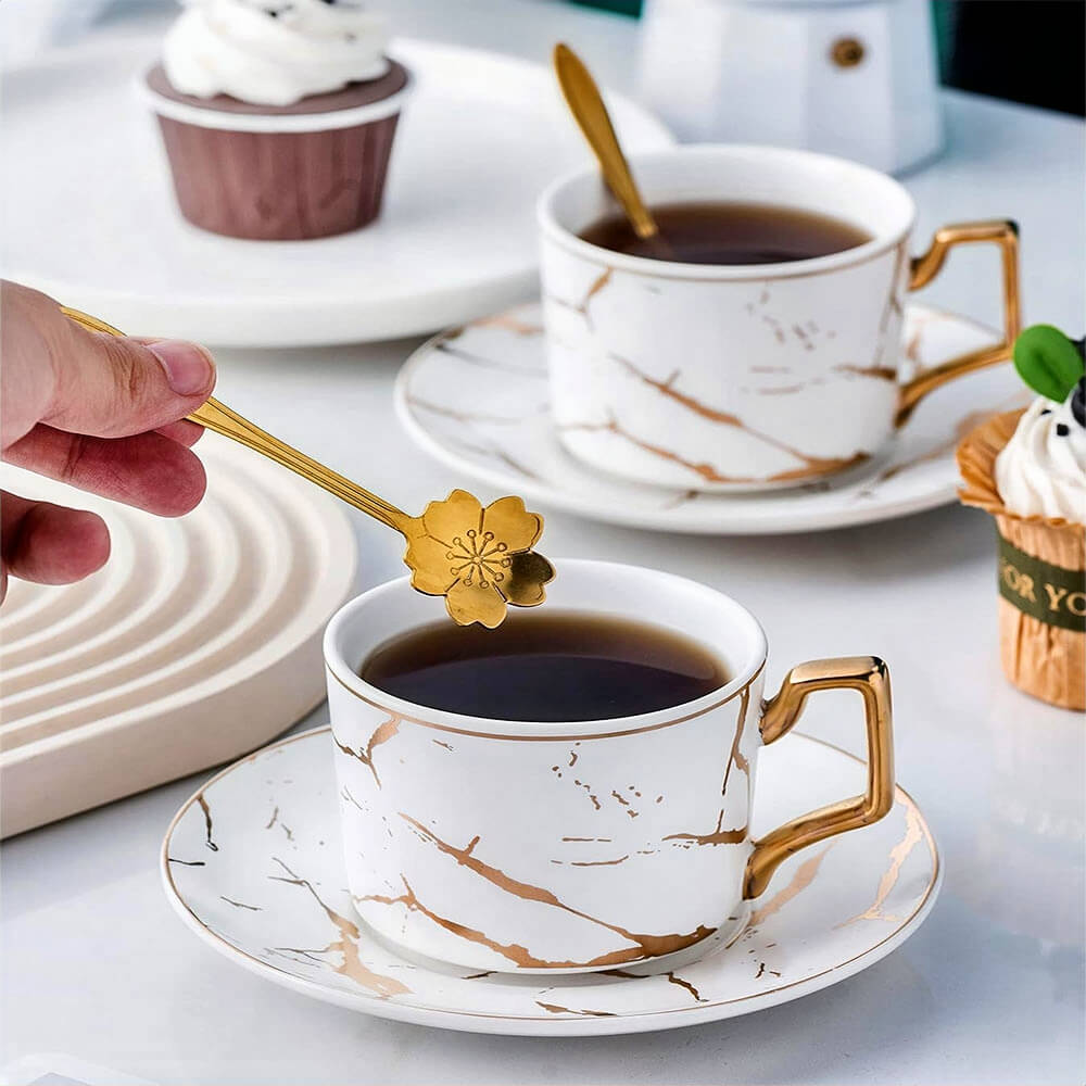 A tea setup featuring two luxurious teacups with matching saucers, designed with a white marble pattern accented by elegant gold veining and golden handles. One cup is being stirred with a delicate gold flower-shaped spoon, while a cupcake and minimalist decor in the background enhance the refined and cozy atmosphere.