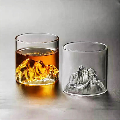 Two glasses with sculpted mountain peaks at their base. One glass is filled with an amber liquid, which highlights the intricate mountain design, while the other glass is empty, revealing the clear and detailed craftsmanship of the glass. The glasses are placed on a smooth surface with a neutral background, emphasizing their unique design.