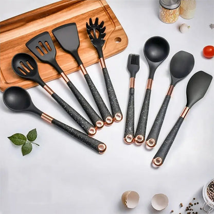 Black silicone non stick utensils with speckled handles and copper accents arranged on a white surface beside a wooden cutting board