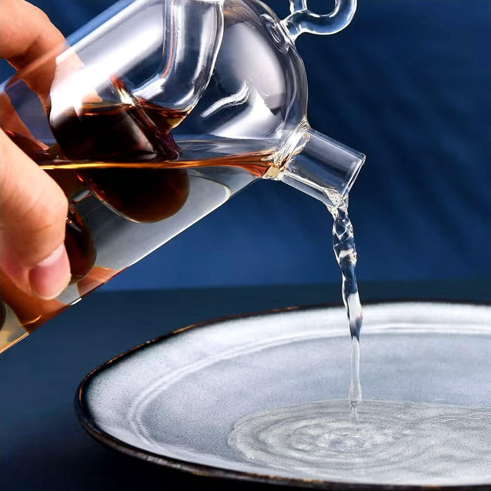 Close-up of a glass oil and vinegar bottle being poured onto a plate. The bottle features a dual-chamber design for storing oil and vinegar separately, with a precise spout for controlled dispensing. The background is dark blue, and the plate has a textured ceramic surface.