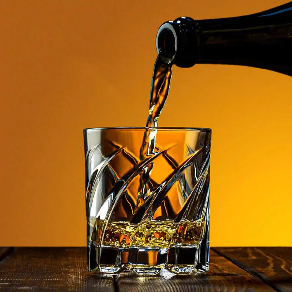 Old fashioned whiskey glasses with a faceted geometric texture as a beverage is poured into the glass on a wooden table.