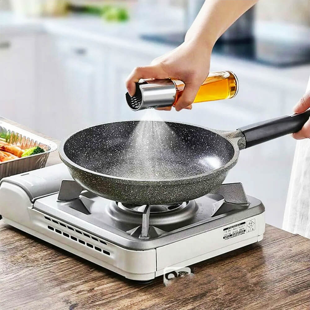 A person using a sleek glass and stainless steel olive oil spray bottle to spray oil evenly onto a non-stick frying pan placed on a portable gas stove. The fine mist covers the pan, ensuring minimal oil usage for healthy cooking. A tray with vegetables is visible in the background, highlighting a clean and organized kitchen setting.