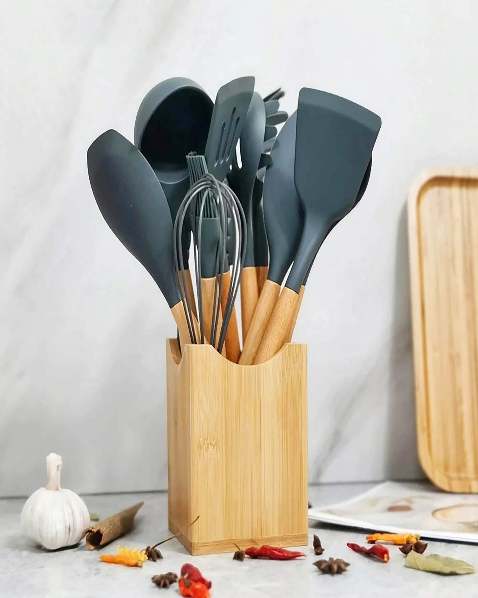 A modern set of OXO kitchen utensils with black silicone heads and wooden handles, neatly organized in a bamboo holder. Surrounded by spices, garlic, and a wooden tray, creating a stylish and functional kitchen setup.